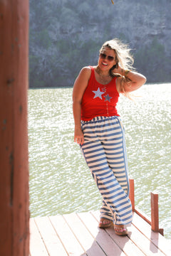 Woman in a red top and blue striped pants standing on a wooden dock by a lake.