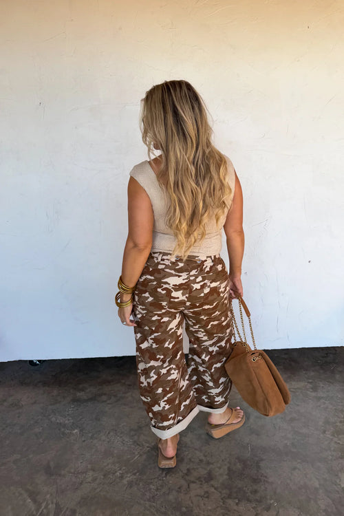 Woman wearing camouflage pants and a beige top, holding a brown bag against a white wall.