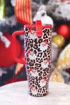 A red handled travel tumbler with a brown leopard print and Santa Claus face pattern, featuring a red lid and straw, set against a blurred Christmas background.