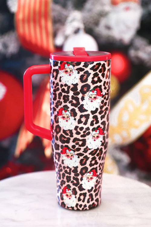 A red handled travel tumbler with a leopard print pattern and Santa Claus illustrations, shown on a light marble surface with a blurred festive background.