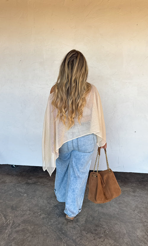 Woman wearing a beige shawl and holding a brown bag against a white wall.