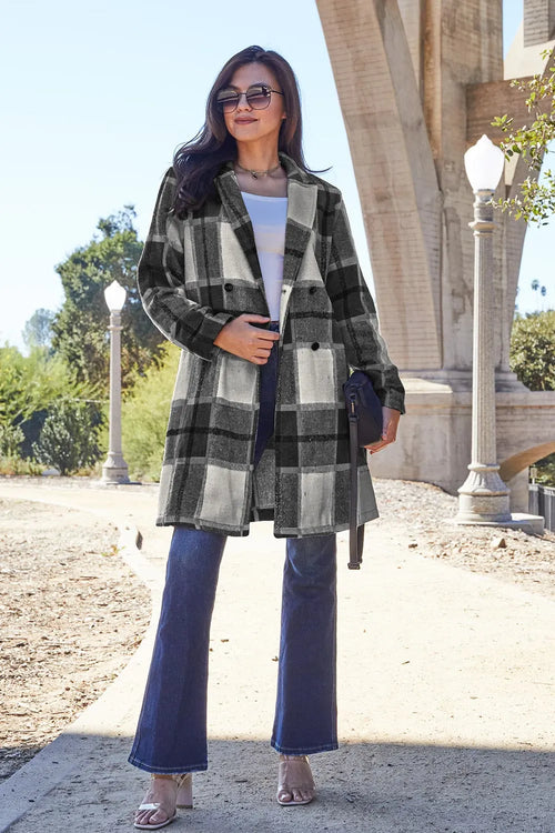 Woman wearing a plaid coat and blue jeans standing outdoors with a building in the background.