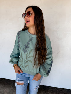 Woman wearing a green sweatshirt with floral designs and ripped jeans against a white wall.