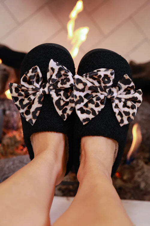 Black slippers with leopard print bows worn by a person in front of a fireplace.
