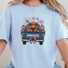 Baby blue graphic tee featuring a highland cow in a vintage truck with America text, fireworks, florals, and American flags.