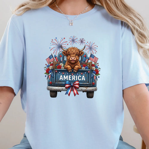 Baby blue graphic tee featuring a highland cow in a vintage truck with America text, fireworks, florals, and American flags.