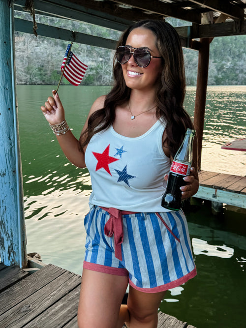 Smiling woman wearing a blue star tank and matching blue denim shorts with red stars, holding a small American flag on a dock.