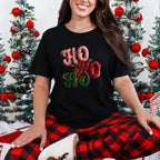 Model wearing black Ho Ho Ho Graphic Tee with red and green holiday decor in background
