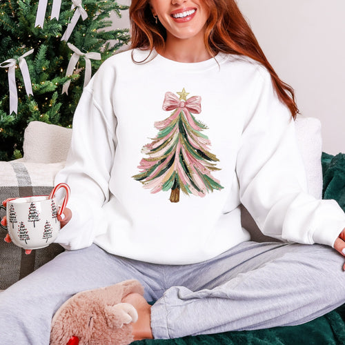 Person wearing a white sweatshirt with a Christmas tree design, holding a mug and a teddy bear, sitting in front of a decorated Christmas tree.