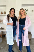 Two women wearing long cardigans over black tops and jeans in a living room setting.