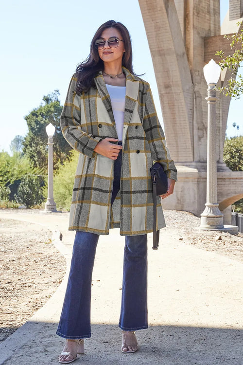 Woman wearing a plaid coat and blue jeans standing outdoors with architectural elements in the background.