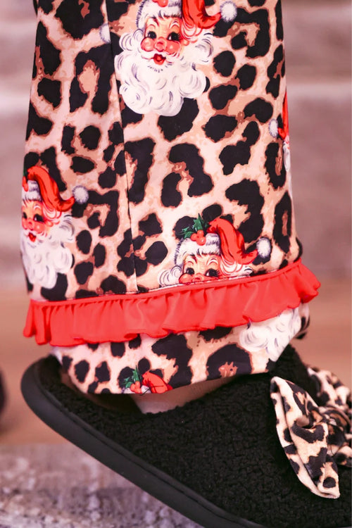 Close-up of a person wearing festive pajama pants with a leopard print pattern, Santa Claus faces, and a red ruffle hem, paired with a black fuzzy slipper featuring a matching leopard print bow.