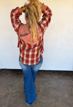A woman viewed from behind wearing a red and white plaid flannel shirt with a peach-colored hood, blue jeans, and gold bracelets, standing against a white wall.