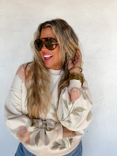 Woman wearing a cream-colored sweater with floral patterns and sunglasses against a white background