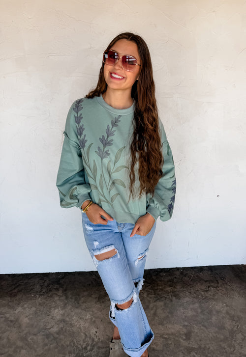 Woman wearing a green sweater with leaf pattern and ripped jeans against a white wall.