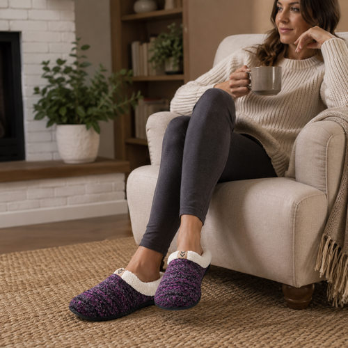 A woman sits in a beige armchair in a cozy living room, wearing a knit purple slipper with a white fuzzy trim and a small gold bow. She is also wearing dark gray leggings and a cream ribbed sweater, holding a gray mug.