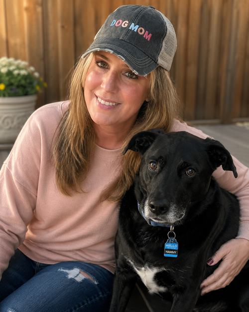 A woman wearing a distressed black 'DOG MOM' trucker hat with colorful lettering, a dusty pink sweatshirt, and blue jeans sits outdoors with a black dog on a patio in front of a wooden fence.