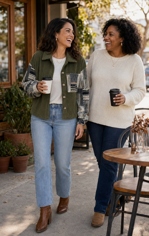 Two women walking and smiling outdoors at a cafe, each holding a black coffee cup. The woman on the left wears an olive green patchwork jacket over a white top and light blue jeans, while the woman on the right wears a cream knit sweater, dark blue jeans, and tan ankle boots.