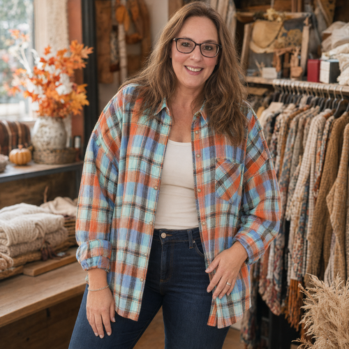A woman wearing a long-sleeve plaid button-up shirt in orange, blue, and light green, layered over a white tank top and dark blue jeans, standing inside a rustic clothing boutique.