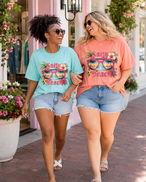 Two women walking together on a sidewalk, wearing matching t-shirts with sunglasses and floral designs.