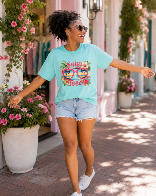 Woman wearing a light blue t-shirt with 'Salty Beach' design, standing outdoors with floral decorations.