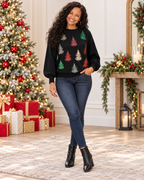 A woman stands in a decorated holiday room wearing a black crewneck sweater with colorful embroidered Christmas tree motifs, dark blue jeans, and black ankle boots. A lit Christmas tree, wrapped gifts, and a mantel are visible in the background.