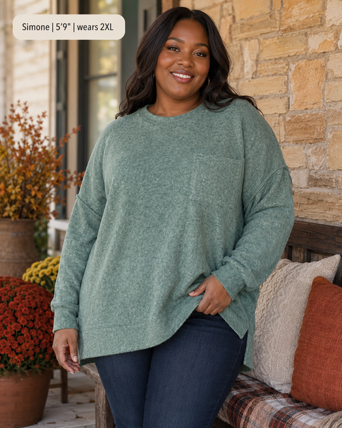A woman modeling an oversized sage green knit sweater with a chest pocket, paired with dark blue jeans, standing on a porch next to a stone wall and decorative pillows.
