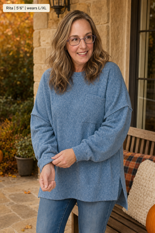 A woman wearing a heathered dusty blue long-sleeve knit sweater with a chest pocket, paired with blue jeans, standing on a stone patio porch.