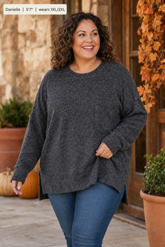 A woman wearing a dark gray heathered knit sweater and blue jeans, standing in front of a rustic stone and wood door with pumpkins and potted plants.
