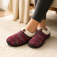 A person wearing cozy knit slip-on slippers with a white sherpa lining, standing on a cream carpet. The slippers feature a marled knit pattern in pink, red, and gray, with a small decorative wooden charm on the side.