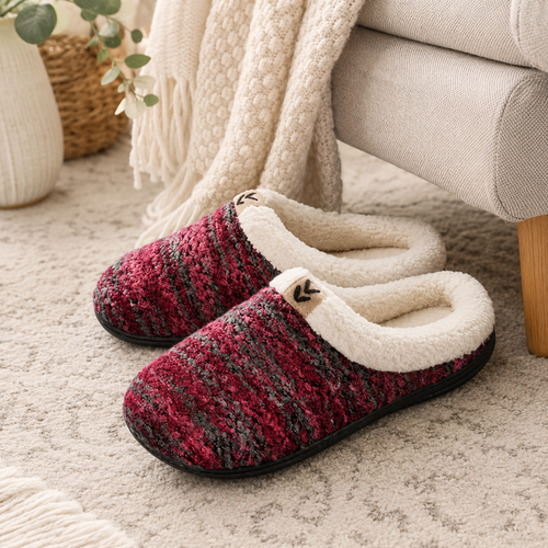 A pair of cozy knit slip-on house slippers with a textured burgundy, red, and gray patterned upper and a soft cream fleece lining, shown on a neutral rug next to a sofa and throw blanket.