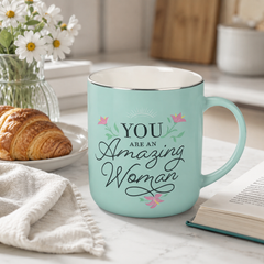 Teal mug with 'You are an Amazing Woman' text on a table with a croissant and flowers.