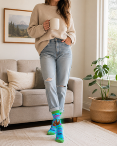 Woman standing in a cozy living room with cuffed jeans and bright blue Sloth Is My Spirit Animal novelty socks featuring green toes and a sloth design.