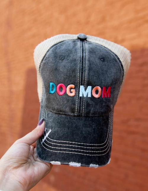 A hand holding a distressed trucker-style baseball cap with a washed black front panel, beige mesh side panels, and colorful embroidered text reading 'DOG MOM' across the front.