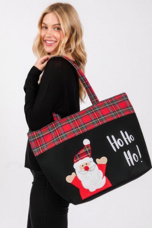 Person holding a black tote bag with red plaid accents and a Santa Claus graphic, on a white background.