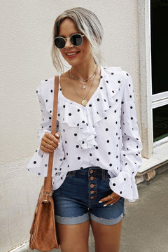 Woman wearing a white blouse with black polka dots and denim shorts, standing against a light-colored wall.