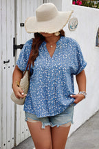 Woman wearing a blue floral blouse and denim shorts with a wide-brimmed hat, standing against a white wall.