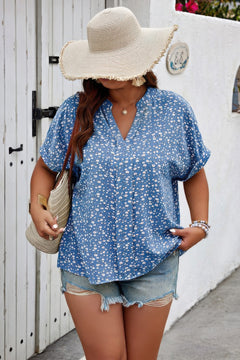 Woman wearing a blue floral blouse and denim shorts with a wide-brimmed hat, standing against a white wall.