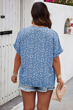 Woman wearing a blue floral blouse and denim shorts, standing in front of a white wall. Back side of blouse.