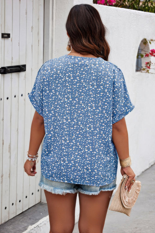 Woman wearing a blue floral blouse and denim shorts, standing in front of a white wall. Back side of blouse.