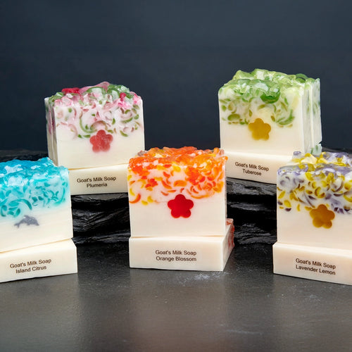 A collection of six handmade goat's milk soap bars displayed on dark slate, featuring colorful textured tops in shades of turquoise, orange, purple, and green with floral and dolphin designs.