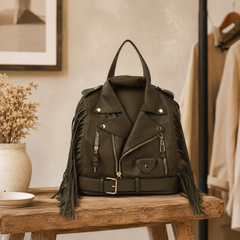An olive green vegan-leather biker jacket backpack with long fringe details and gold-tone hardware, displayed on a rustic wooden bench in a warm, styled interior setting.