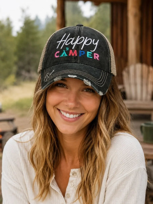 A woman smiling while wearing a distressed black trucker-style cap with 'Happy Camper' embroidered on the front, set against a rustic outdoor background.