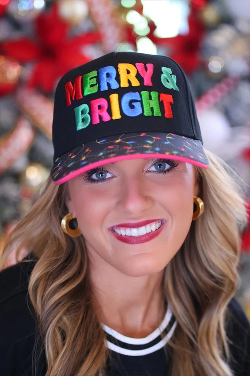 A smiling woman wearing a black baseball cap with colorful 'MERRY & BRIGHT' lettering and a pink brim, photographed against a blurred festive holiday background.