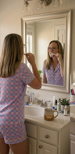 A realistic morning scene of a woman with straight blonde hair and glasses wearing a purple checkered short pajama set while brushing her teeth in a bathroom mirror. A steaming ceramic mug featuring the "BeYOUtifully Comfortable" logo sits on the white vanity among skincare bottles.