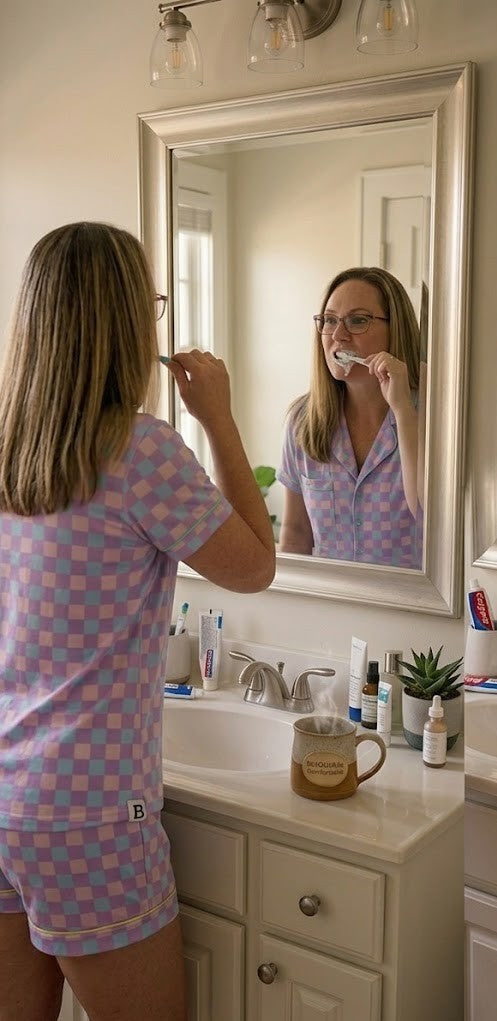 A realistic morning scene of a woman with straight blonde hair and glasses wearing a purple checkered short pajama set while brushing her teeth in a bathroom mirror. A steaming ceramic mug featuring the "BeYOUtifully Comfortable" logo sits on the white vanity among skincare bottles.