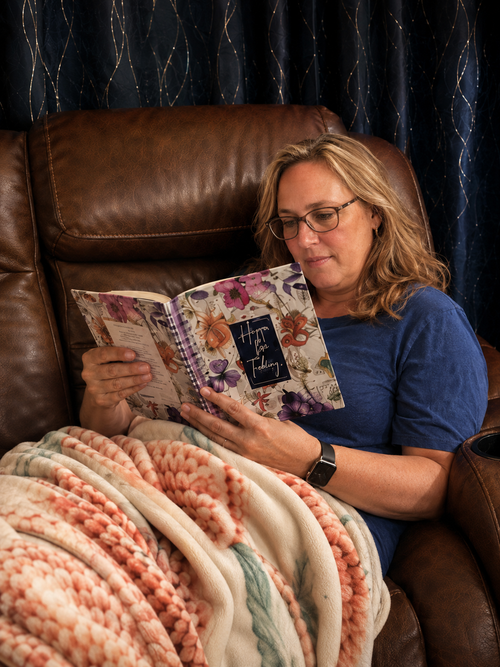 Rita relaxing on a brown leather couch under a cozy blanket while reading Hope for Today, a floral-covered devotional book.