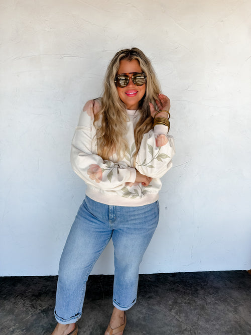Woman wearing a cream sweater and blue jeans against a white wall.