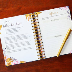Open planner with floral design on a wooden surface, featuring a pen.