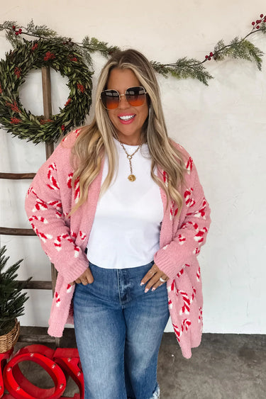 Woman wearing a pink cardigan with candy cane pattern, white shirt, and blue jeans in front of a Christmas wreath.